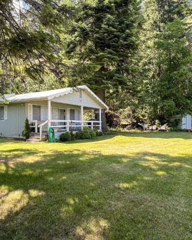 Single-story cabin with covered porch surrounded by tall trees and sunny lawn