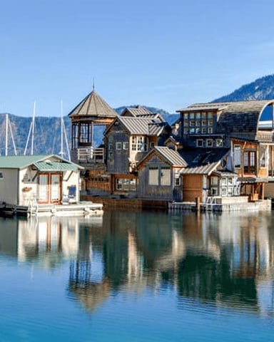 Waterfront marina with sailboats and colorful boathouses reflected in calm water, forested mountains in background