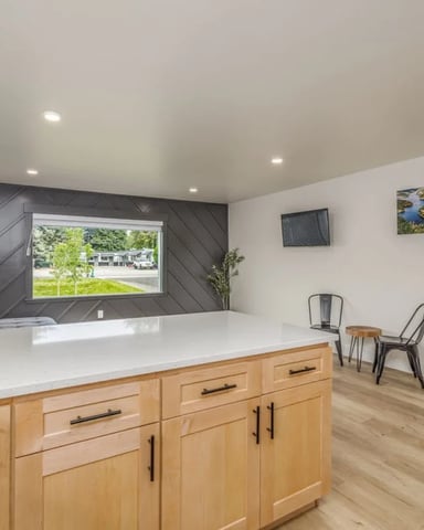 Modern open-plan living space with wood cabinetry, white countertop, black accent wall, built-in bed, and large window overlooking yard