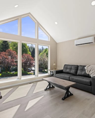 Modern living room with black leather sofa, large windows overlooking landscaped yard with flowering trees, vaulted ceiling with skylights, and wood flooring.